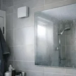 Après la douche, le miroir reste légèrement embué et des gouttelettes se déposent sur la surface. On distingue en partie haute un petit extracteur mural de ventilation mécanique répartie (VMR), posé discrètement pour aider la pièce à évacuer l’humidité. La lumière froide du matin et la serviette humide sur la porte donnent une scène très concrète de ce que vous vivez au quotidien quand la ventilation doit vraiment faire son travail.